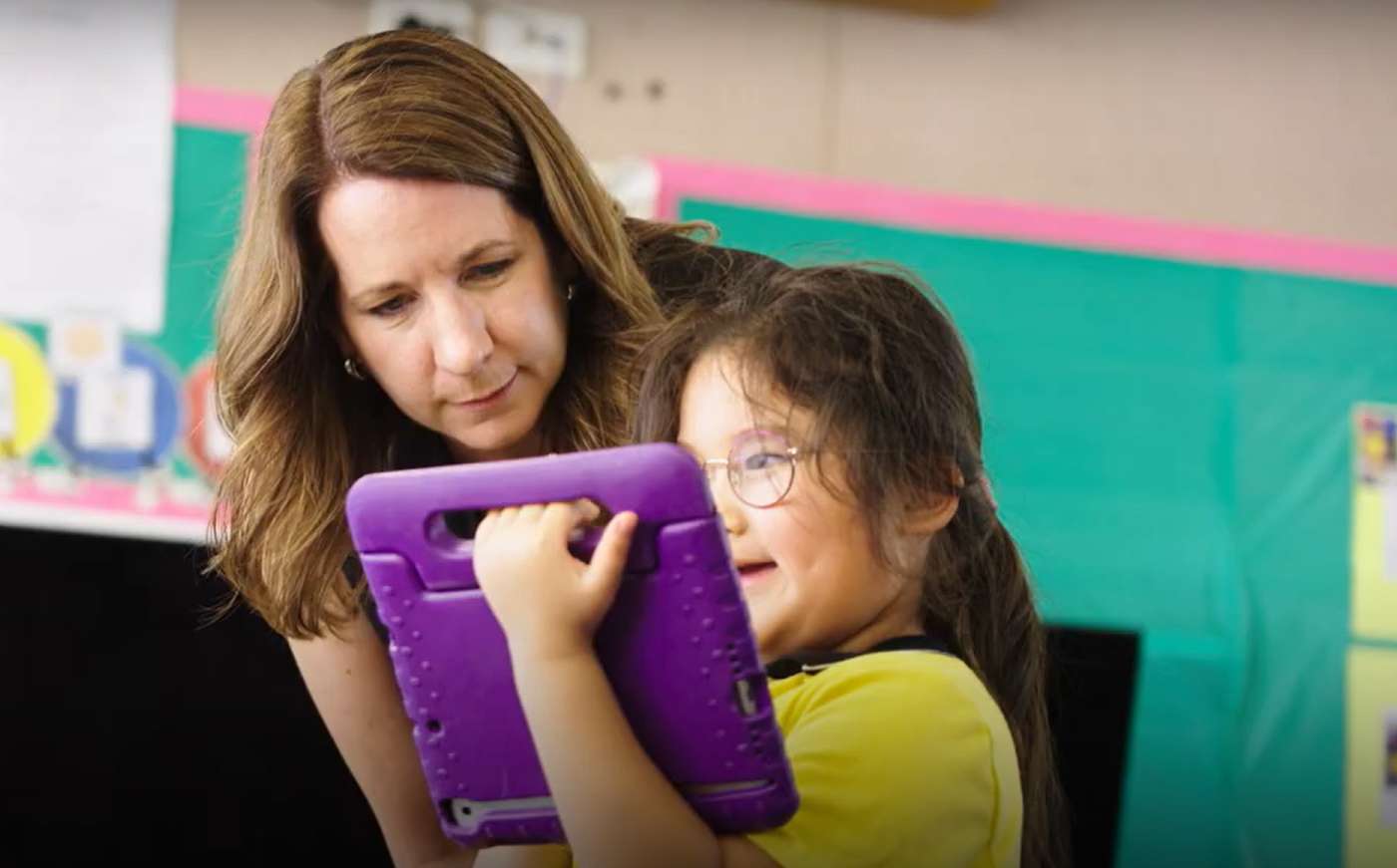 A child is holding a tablet while teacher and child are looking at it