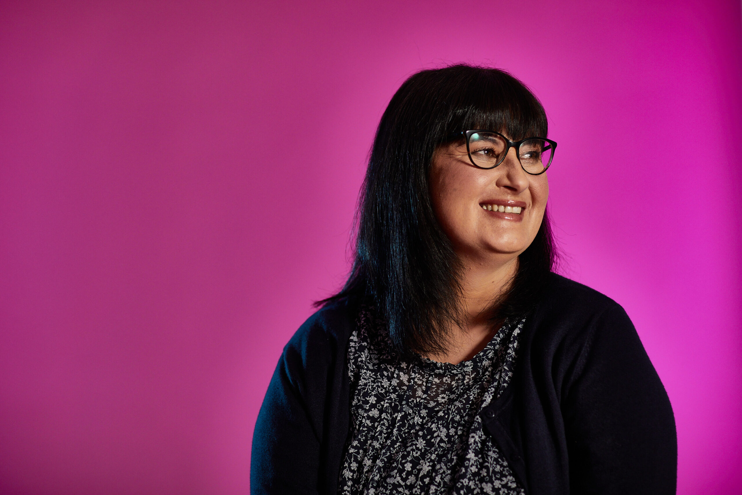 Woman wearing glasses smiling sitting in front of a plum coloured background
