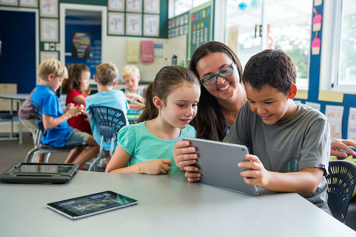 Teacher and children working on digital devices in busy classroom